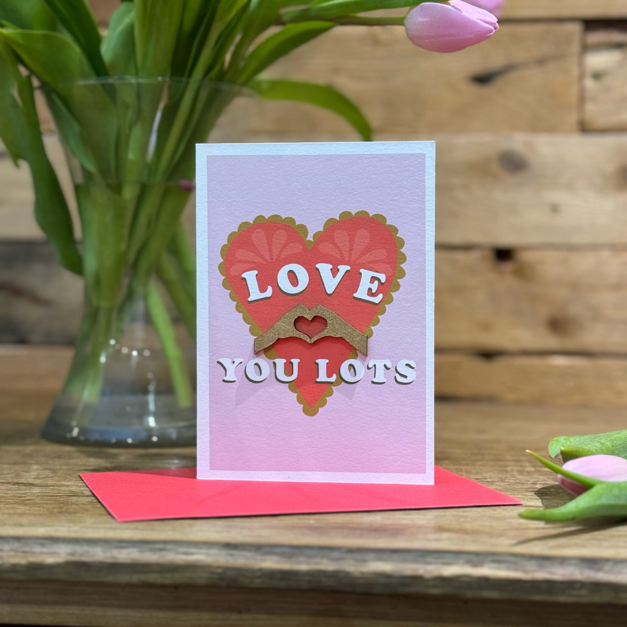 Card with pink background and red heartt with gold 3D hands making a heart shape