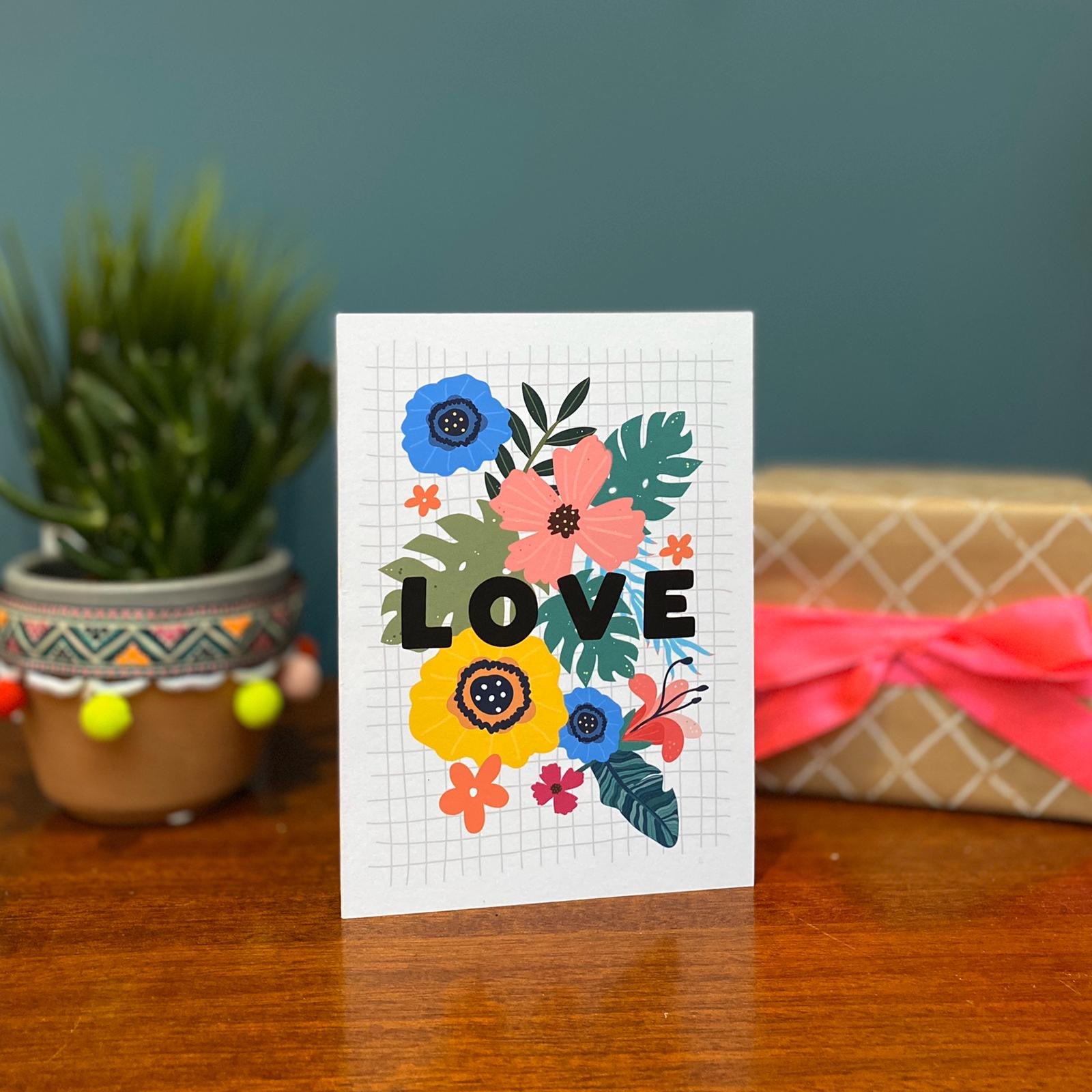 A hand with dark nail polish holds a Bright Floral Love Card by Hendog Designs, beautifully designed on A6 cardstock. In the background, a vase overflows with red, purple, yellow, and orange tulips set against a blue wall.