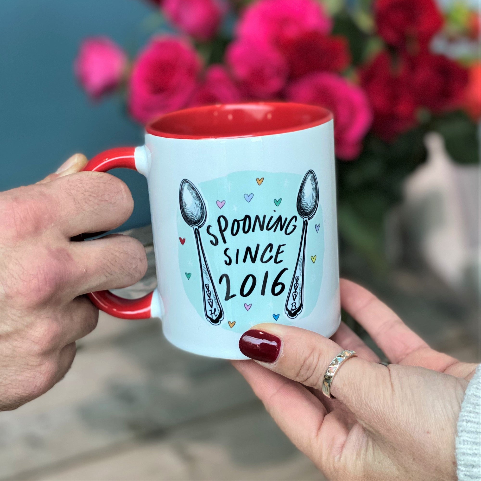 Person handing a red and White china mug to someone with a "Spooning Since 2016" on the front and Spoons Illustrations 