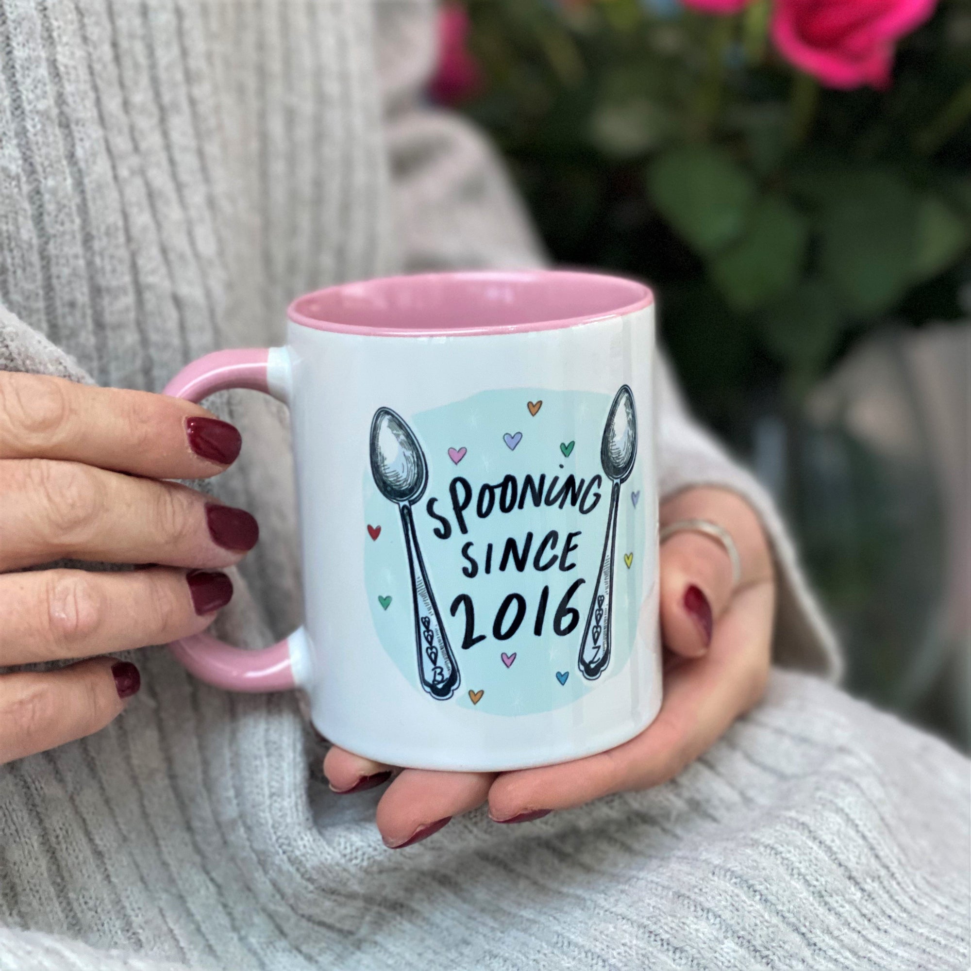 Person holding a white and pink mug with "Spooning Since 2016" and spoon illustrations. Red roses in the background.