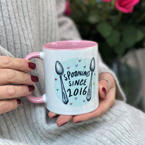 Person holding a white and pink mug with "Spooning Since 2016" and spoon illustrations. Red roses in the background.