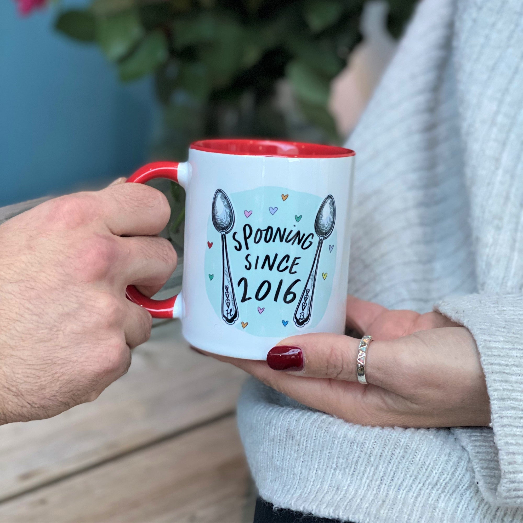 Person giving a person a mug. The china mug is Red and white with "spooning since 2016" design on the front