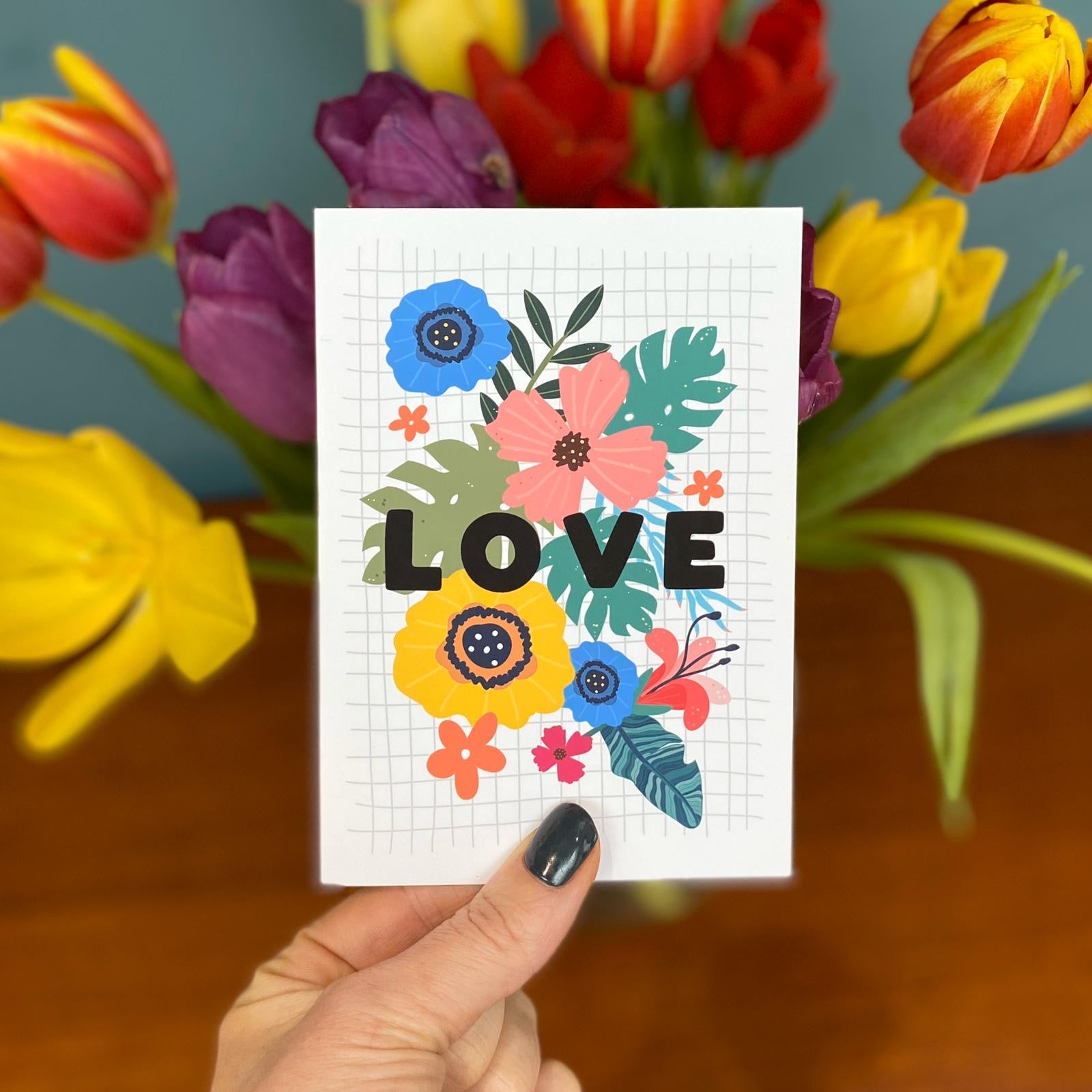 A hand with dark nail polish holds a Bright Floral Love Card by Hendog Designs, beautifully designed on A6 cardstock. In the background, a vase overflows with red, purple, yellow, and orange tulips set against a blue wall.