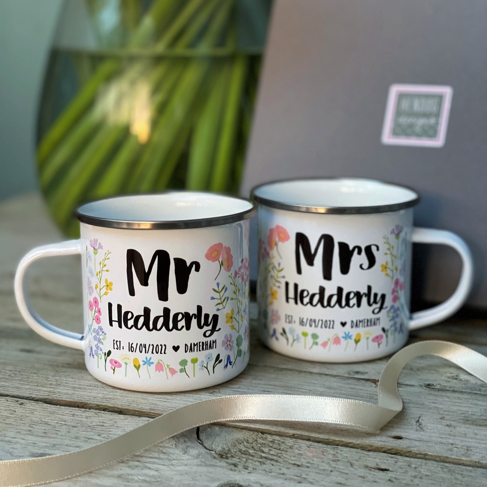 The Couples Floral Personalised Wedding/Anniversary Enamel Mug Set by Hendog Designs features two mugs with “Mr Hedderly” and “Mrs Hedderly,” floral designs, and the date 16/07/2022. Perfect as a wedding gift, theyre presented with a ribbon and a blurred gift box in the background.