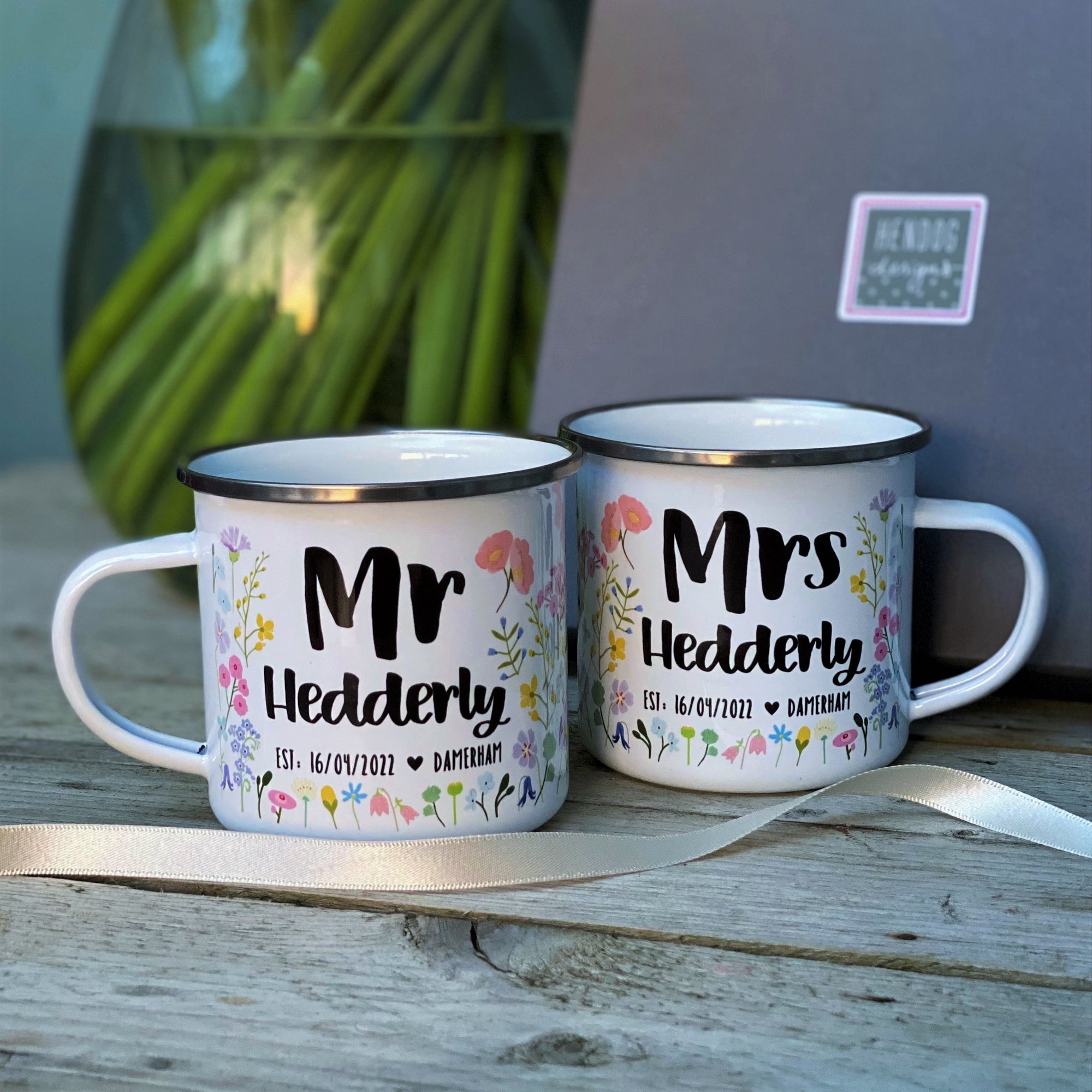 Perfect for a wedding gift, the Couples Floral Personalised Wedding/Anniversary Enamel Mug Set by Hendog Designs includes two white mugs reading Mr Hedderly and Mrs Hedderly, with Est. 16/07/2017 and Darterham. Resting on wood beside a gray box and green plant stems, they add charm to any setting.