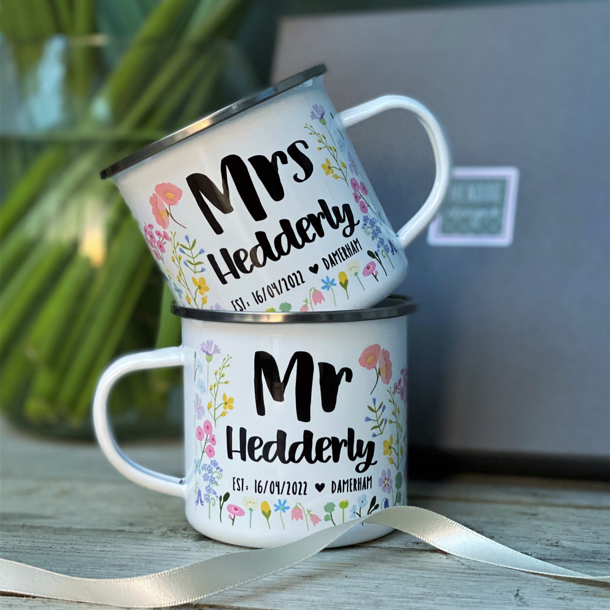 The Couples Floral Personalised Wedding/Anniversary Enamel Mug Set by Hendog Designs features two white enamel mugs stacked elegantly. The top reads Mrs Hedderly, the bottom Mr Hedderly, with Est. 16/01/2022 and a heart symbol, making it an ideal personalised wedding gift. A light ribbon completes the look.