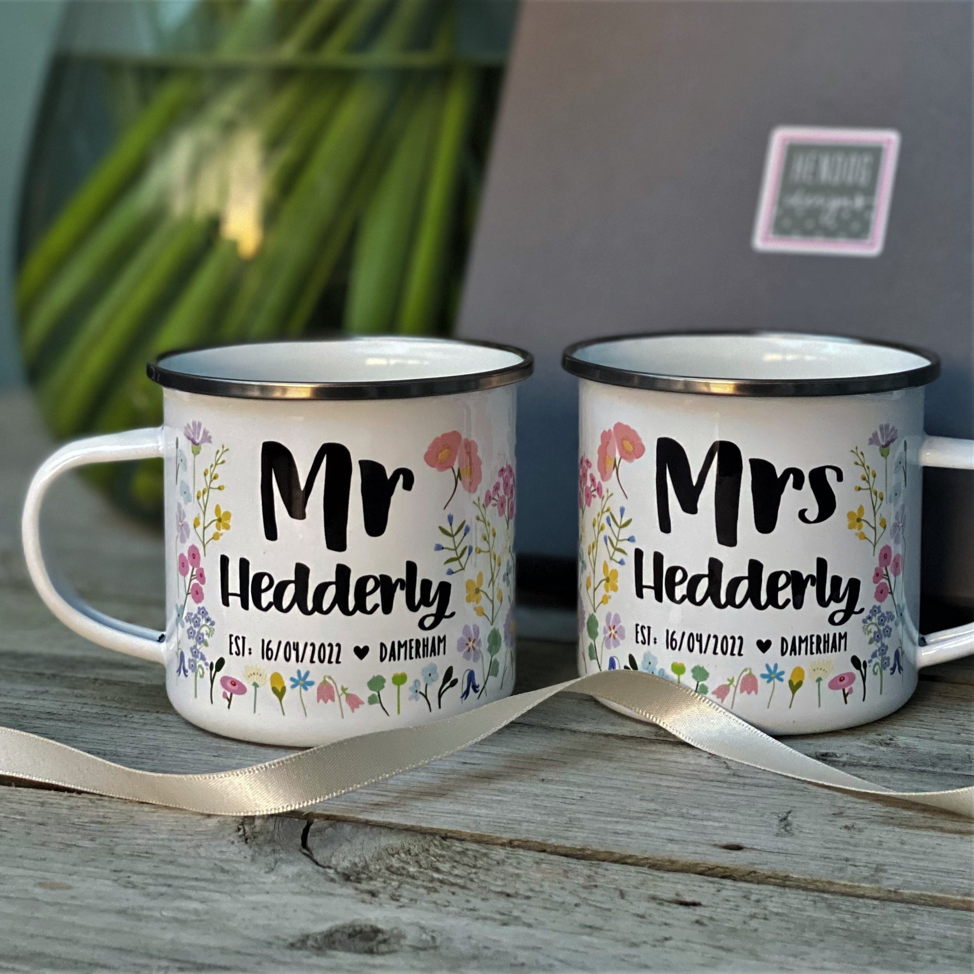 The Couples Floral Personalised Wedding/Anniversary Enamel Mug Set by Hendog Designs features two mugs with Mr Hedderly and Mrs Hedderly text, colorful floral designs, and the date Est. 16/07/2022, tied with a ribbon on a wooden surface near a laptop and vase. Charming wedding gifts!.