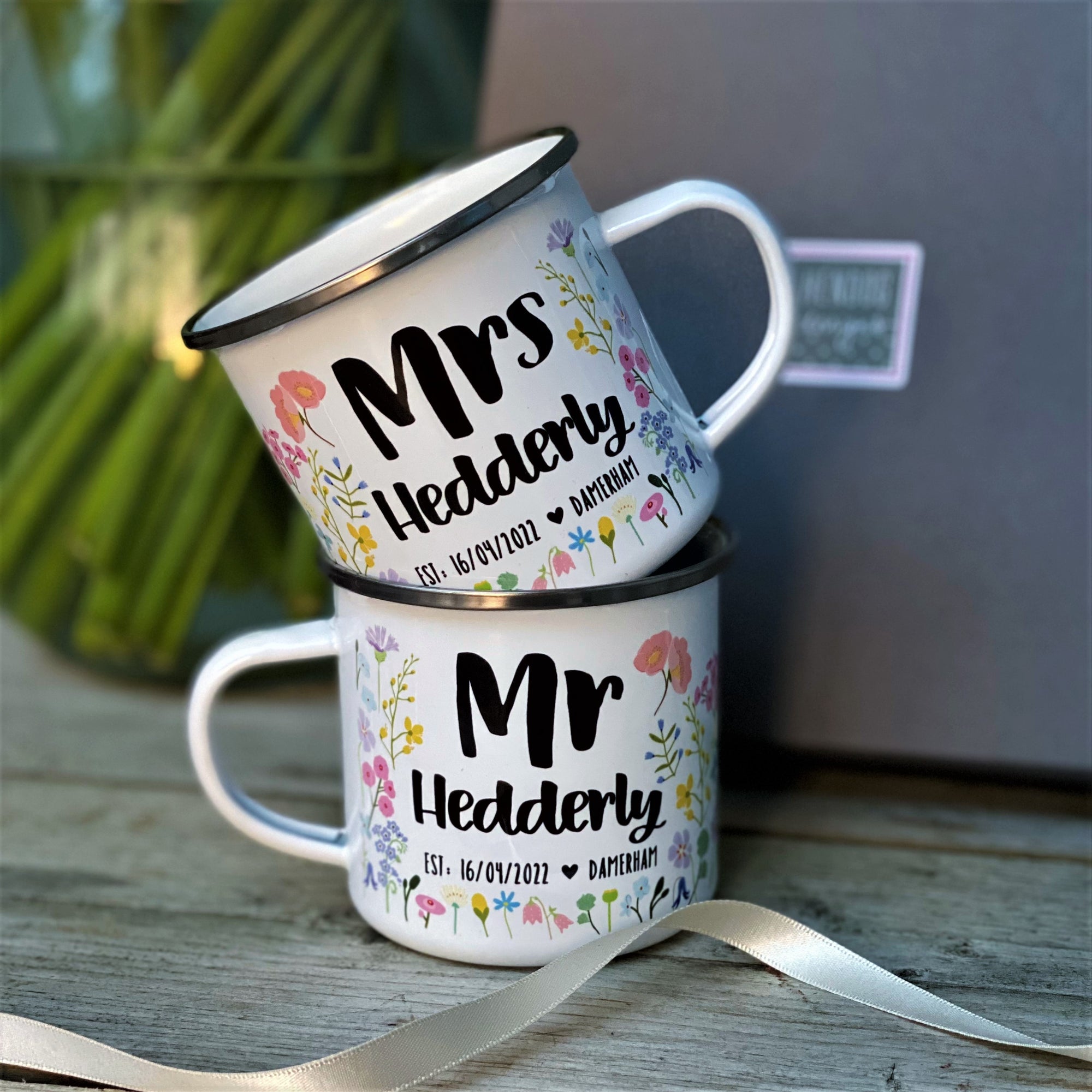 The Couples Floral Personalised Wedding/Anniversary Enamel Mug Set by Hendog Designs features two stacked mugs reading Mrs Hedderly and Mr Hedderly with dates, complemented by a ribbon on a wooden surface—a charming gift.